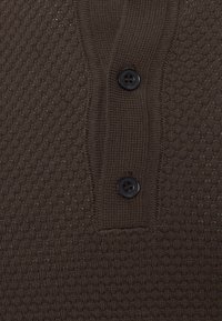 Hnědá polo košile s texturou, která má diamantový vzor, tři černé knoflíky a pletenou tkaninu s jemným detailem.