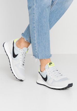 Zapatillas Nike en gris y negro, con parte superior de malla y gamuza, detalles en amarillo neón y suela de goma texturizada.