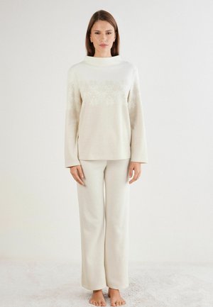 Creme gebreide trui met een hoge kraag, gekenmerkt door een sneeuwvlokpatroon, gecombineerd met bijpassende getextureerde broek met wijde pijpen. Zachte, comfortabele stof.