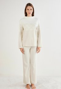 Creme gebreide trui met een hoge kraag, gekenmerkt door een sneeuwvlokpatroon, gecombineerd met bijpassende getextureerde broek met wijde pijpen. Zachte, comfortabele stof.