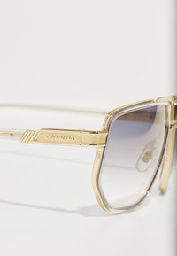 Gafas de sol con montura dorada, varillas transparentes y lentes gris degradado. Presentan una marca en la varilla y acentos texturizados en la montura.