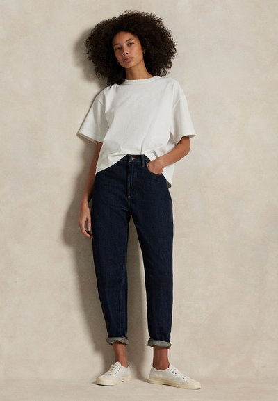 Kobieta o kręconych włosach, ubrana w luźną białą koszulkę, ciemne podwinięte dżinsy i białe trampki, stojąca na tle beżowej ściany.