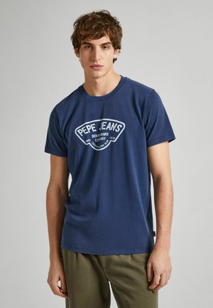 Camiseta de algodón azul marino con mangas cortas y cuello redondo, con un logo blanco impreso en el centro. El modelo lleva pantalones verdes oliva.