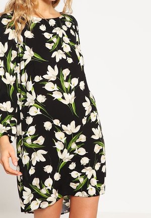 Woman wearing a black long-sleeve dress with a white and green floral pattern, standing against a plain background.