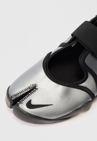 Ezüst színű sportcipő texturált felső résszel, fekete részletekkel és elasztikus pánttal. Jellegzetes varrási dizájnnal és Nike logóval a lábujjaknál.