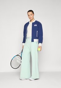 Navy bomber jacket with white accents, light green wide-leg pants, and white sneakers. Holding a tennis racquet and a yellow tennis ball.