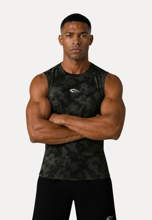 Muskelöser Mann steht mit verschränkten Armen da und trägt ein schwarzes ärmelloses Camouflagetop sowie schwarze Sportschorts vor einfachem Hintergrund.