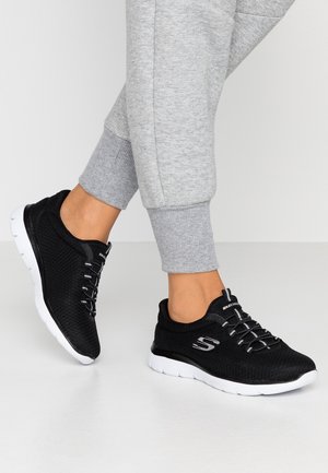 Osoba nosząca czarne sneakersy Skechers z białymi podeszwami i szare spodnie dresowe, stojąca na białej podłodze na jednolitym tle.