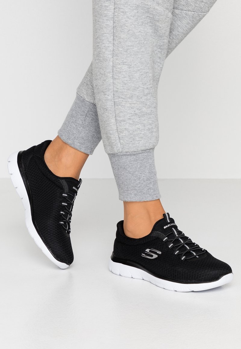 Personne portant des baskets Skechers noires à semelles blanches et un pantalon de survêtement gris, debout sur un sol blanc devant un fond uni.