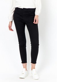 Jeans negros ajustados con una cintura de altura media, que cuentan con un dobladillo deshilachado y una textura suave, combinados con zapatillas blancas sobre un fondo neutral.