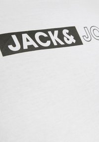 Camiseta blanca con un estampado de rectángulo negro que presenta el texto "JACK&" en negrita y "JO" en una fuente más delgada, mostrando una textura suave.