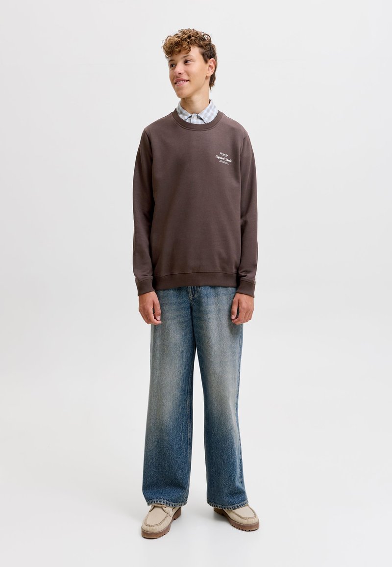 Jeune personne aux cheveux bouclés portant un sweat-shirt marron, une chemise à carreaux claire, un jean large bleu et des chaussures beige, debout sur un fond blanc.