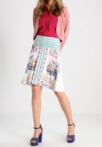 Jupe à motif floral avec une base blanche, une ceinture verte et des motifs colorés. Assortie avec un cardigan rose et des sandales à plateforme bleues.