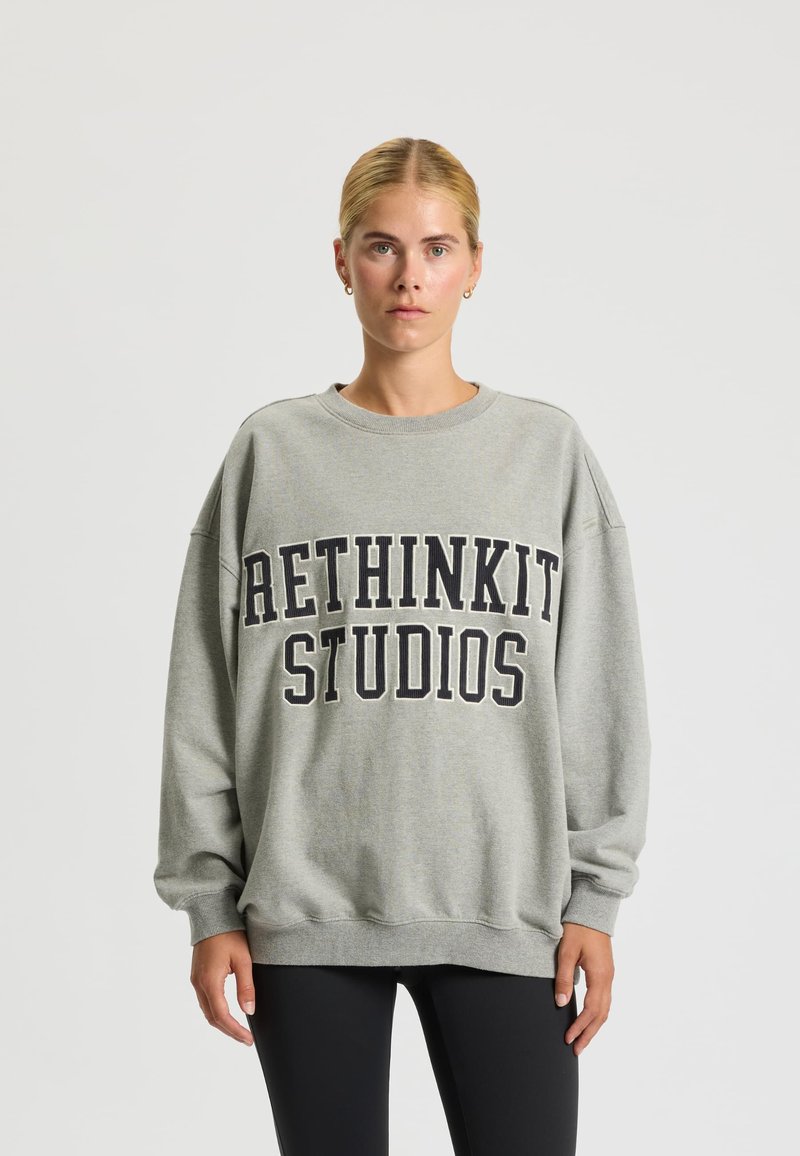 Felpa grigia oversize con "RETHINKIT STUDIOS" stampato in grassetto nero sul petto. Tessuto morbido e polsini a coste.