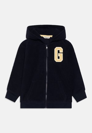 Veste polaire à capuche bleu marine avec fermeture éclair à l'avant, poignets côtelés, poches et un grand écusson beige "G" sur la poitrine gauche.