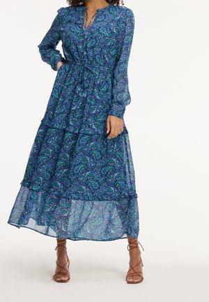 Femme portant une robe maxi à manches longues imprimée paisley bleu et vert avec une ceinture à la taille, associée à des sandales marron à talons hauts et bretelles.