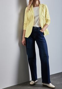 Gele blazer over een wit shirt, gecombineerd met donkerblauwe wijde jeans en witte sneakers. De blazer heeft een enkele knoop en zakken.