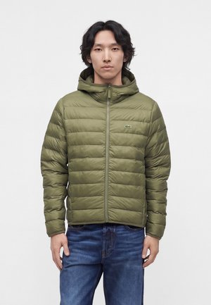 Uomo che indossa una giacca imbottita verde oliva con cappuccio e zip e jeans blu, in piedi davanti a uno sfondo bianco uniforme.