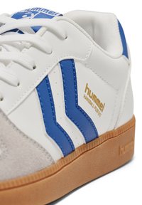 Witte sneaker met blauwe strepen, suède accenten en een zool van natuurlijk rubber. Gouden logotekst "hummel HANDBALL PERFECT" aan de zijkant. Ontwerp met afgeronde neus.