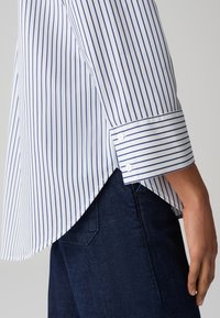Camicia con bottoni, caratterizzata da strisce verticali blu su uno sfondo bianco. Presenta un colletto a punta, polsini con chiusura a bottone e un orlo leggermente più lungo sul retro.