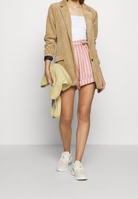 Beige corduroy blazer, witte textuur top, roze en witte gestreepte shorts, houdt een lichtgele jas vast, gecombineerd met witte sneakers.