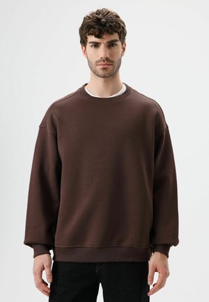 Sweat-shirt marron à col rond, coupe ample et poignets côtelés. Le tissu a une texture lisse, avec des détails de couture subtils. Associé à un pantalon foncé.