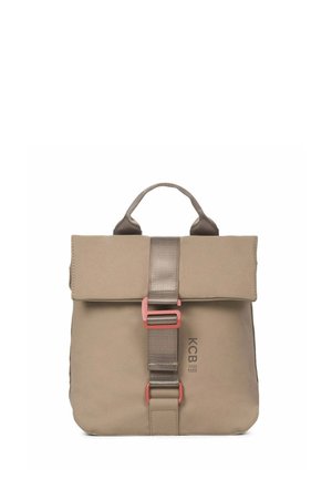 Vrouw draagt een lichtblauwe trui, losse blauwe spijkerbroek, witte sneakers en een beige rugzak met een voorste gesp, staand tegen een effen achtergrond.