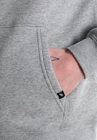 Ruka částečně v přední kapse šedého mikinového svršku Nike, viditelný malý černý štítek Nike a jednoduchá linková tetování na zápěstí.