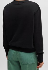 Osoba v černém dlouhém rukávu a zelených kalhotách, zobrazená zezadu na jednoduchém šedém pozadí.