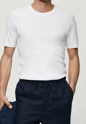 Mann trägt ein eng anliegendes weißes kurzärmeliges, strukturiertes Strickshirt und marineblaue Hosen mit Kordelzug, hält marineblauen Stoff in der linken Hand.