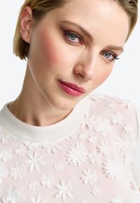 Weißer Sweatshirt mit floralem Design, das eine strukturierte Oberfläche mit unterschiedlich großen Blumen und weißen Perlenakzenten aufweist, ergänzt durch einen gerippten Rundhalsausschnitt.