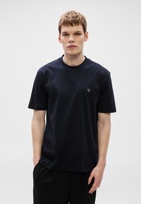 Μπλε ναυτικό βαμβα�κερό μπλουζάκι με κοντά μανίκια και λαιμόκοψη στρογγυλή, με ένα μικρό στρογγυλό λογότυπο στο στήθος. Λείο ύφασμα, αργκό κοψίματος.