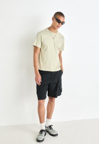 T-shirt vert clair à manches courtes associé à un short cargo noir. Des lunettes de soleil noires, un collier en chaîne argenté et des chaussures multicolores complètent la tenue.