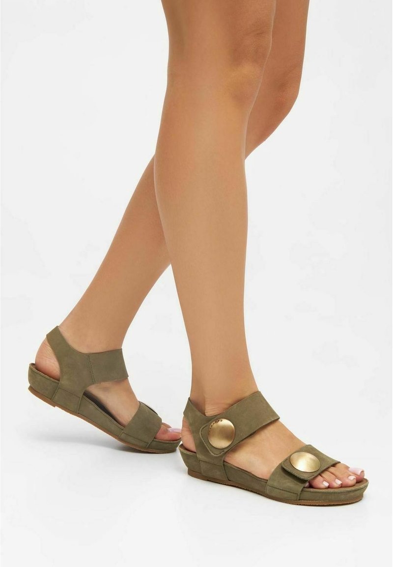 Jambes féminines portant des sandales ouvertes vert olive avec de larges brides et de grandes boucles rondes dorées, marchant sur une surface blanche.