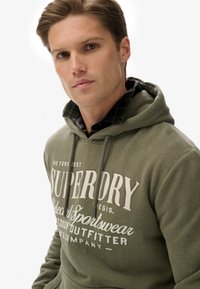Sudadera de color verde oliva con un bolsillo frontal y texto blanco. Presenta cordones en la capucha y está combinada con una camisa de cuadros oscura debajo.