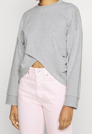 Personne portant un haut gris clair à manches longues avec un ourlet croisé et un pantalon rose pâle taille haute, les bras détendus le long du corps.