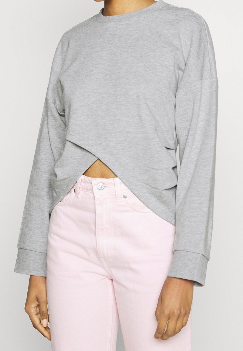 Personne portant un haut gris clair à manches longues avec un ourlet croisé et un pantalon rose pâle taille haute, les bras détendus le long du corps.
