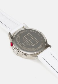 Silver stainless steel watch with a white textured strap. Features a round case, a gray back with "TOMMY HILFIGER," and a red crown accent.