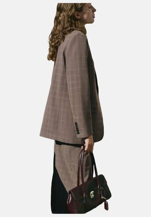 Blazer de cuadros marrón con hombros estructurados, combinado con una falda a juego. Modelo sosteniendo un bolso de piel burdeos con asas para el hombro.