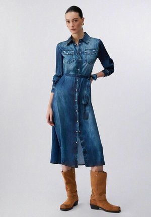 Donna che indossa un vestito lungo in denim blu con maniche lunghe, abbottonato sul davanti e con cintura, e stivali di camoscio marrone a metà polpaccio, in piedi con una mano sul fianco.