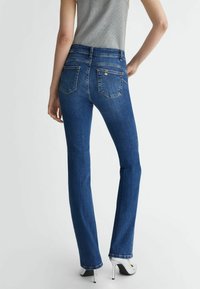 Blauwe denim flared jeans met een hoge taille, gedetailleerde stiksels en twee achterzakken. Draagt een paar zilveren hoge hakken.