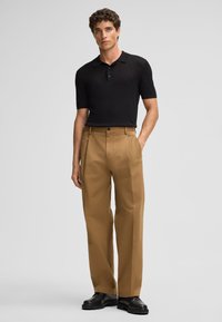 Polo shirt nero a maniche corte, pantaloni khaki con pieghe e scarpe nere. Tessuto liscio, vestibilità su misura e dettaglio con bottone in vita.