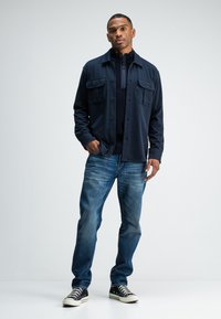 Mann in dunkelblauem Jacket, schwarzem Zip-Neck-Pullover, blauen Jeans und schwarzen High-Top-Sneakern, der mit einer Hand in der Tasche steht.