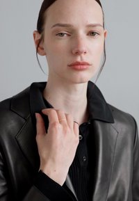 Schwarze Lederjacke über einem schwarzen gerippten Poloshirt; das Modell trägt einen goldenen Ring am Finger, hat glatte Haut und neutral beleuchtete Umgebung.