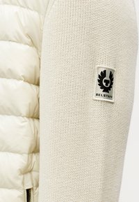 Μπεζ μπουφάν κοντού τύπου με υφασμένα π λεκτά μανίκια. Διαθέτει λογότυπο σε κονκάρδα, συνδυάζοντας γυαλιστερό και ματ φινίρισμα σε ένα ελαφρύ σχέδιο.