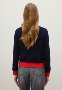 Maglione blu navy con accenti rossi all'orlo e ai polsini, realizzato in materiale morbido, design aderente. Jeans grigi con tasche posteriori visibili.