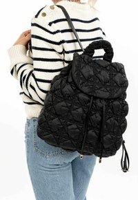 Sac à dos matelassé noir avec un rabat repliable, motif texturé, fermeture à cordon et doubles poignées supérieures. Porté sur un pull à rayures.
