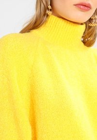 Yellow high-neck sweater with a textured, soft finish. Features a ribbed collar and a relaxed fit. Earrings are gold-toned with round accents.