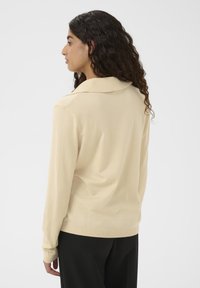 Kvinde med langt krøllet hår iført en beige langærmet sweater og sorte bukser, der står til venstre mod en enkel hvid baggrund.