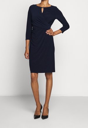 Woman wearing a knee-length, navy blue wrap dress with three-quarter sleeves and black high heels, standing with one hand on hip.